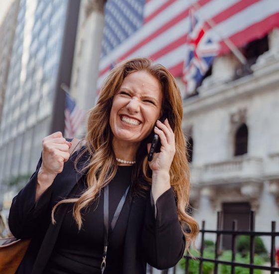 zipwp-image-13801635 Excited Woman Using Mobile Phone on a Street and US Flag in Background